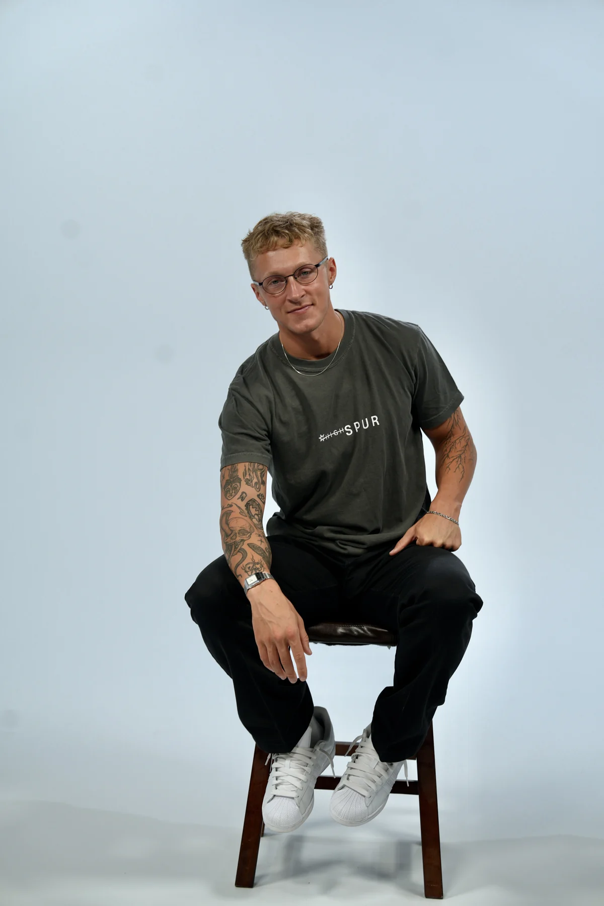 Seated on a wooden stool in a grey HIGHSPUR tee, against a pale blue studio backdrop.